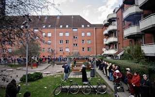 People standing in a courtyard between two apartment buildings while a woman wearing a hijab speaks into a microphone.