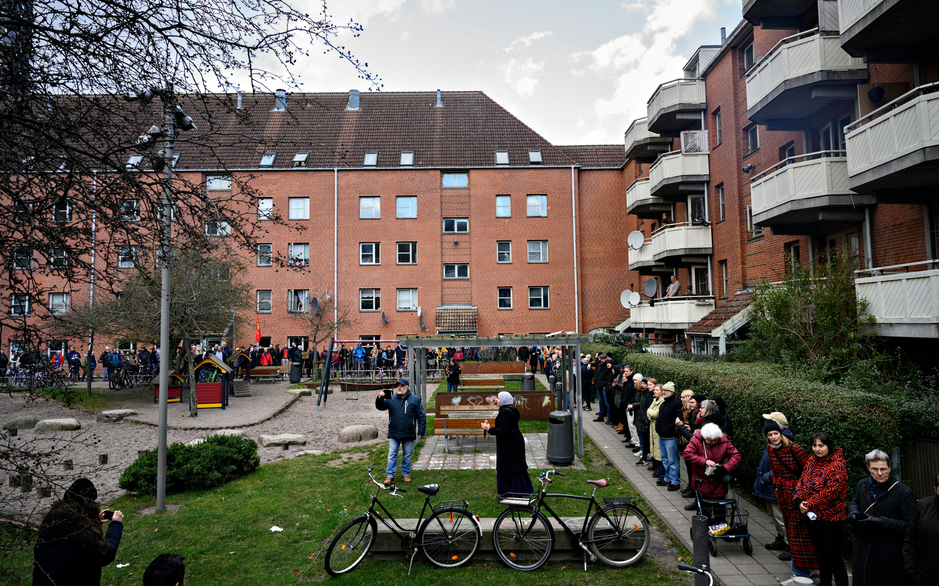 People standing in a courtyard between two apartment buildings while a woman wearing a hijab speaks into a microphone.