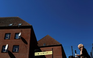 A woman in a pink headscarf walks past red brick buildings under a blue sky